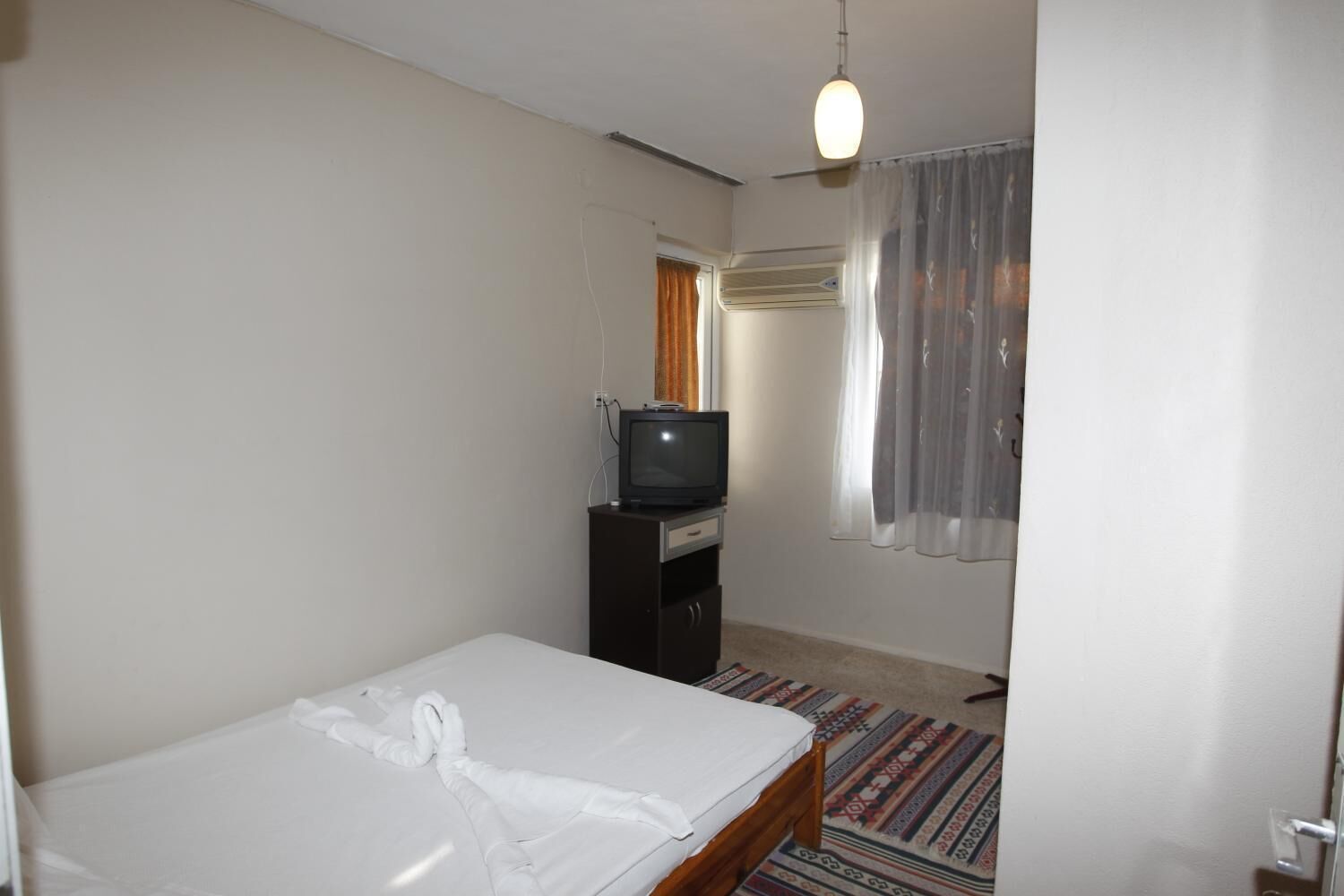 A small, simple hotel room with a single bed, a television on a dresser, and a window with sheer curtains.