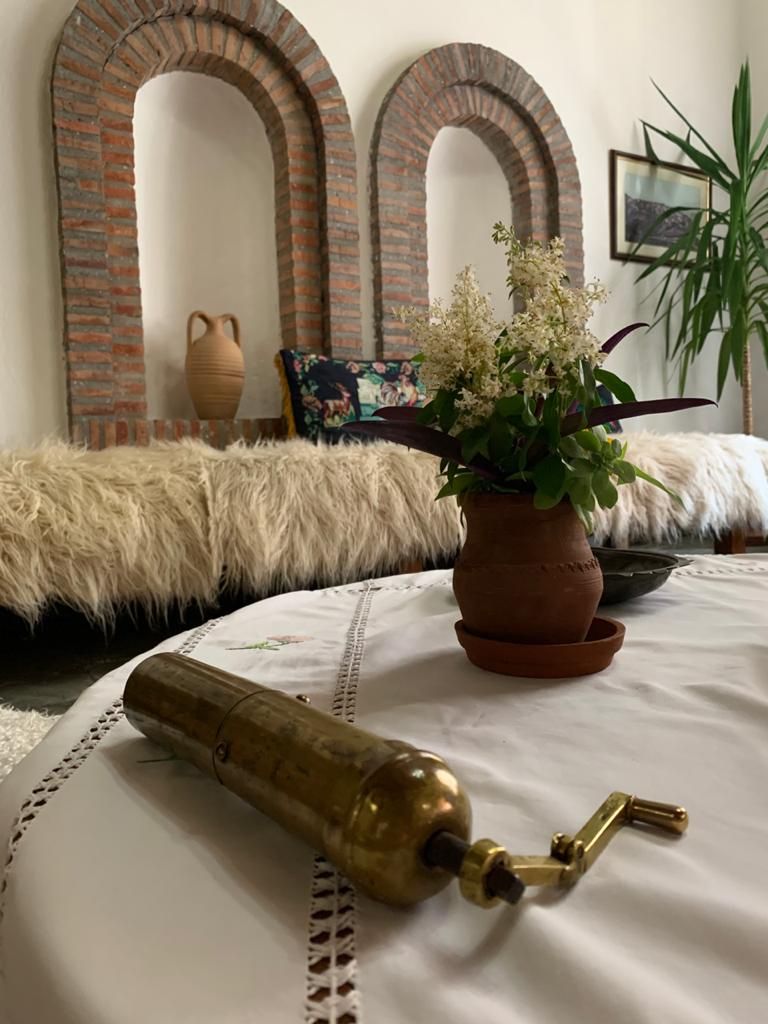 A room with a round table, a brass object, a potted plant, and a brick wall with arches.
