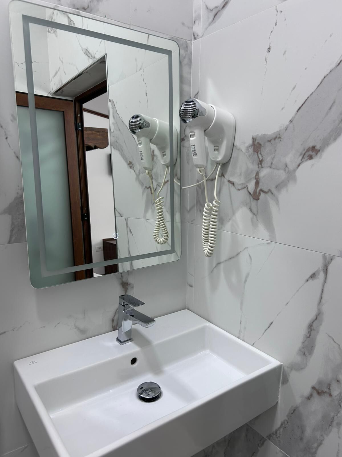 Un baño con pared de mármol, un lavamanos con grifo cromado y dos secadores de pelo montados en la pared.