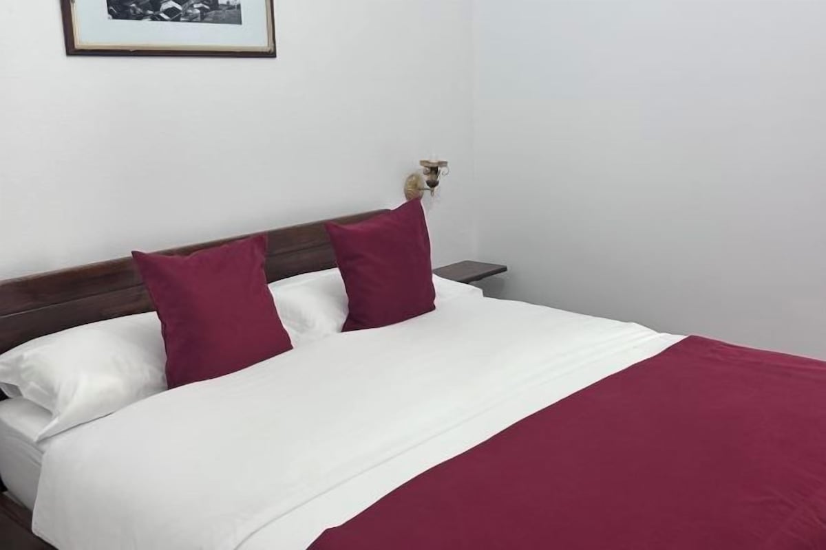 A bed with white and maroon bedding, a wooden headboard, and a nightstand with a lamp.