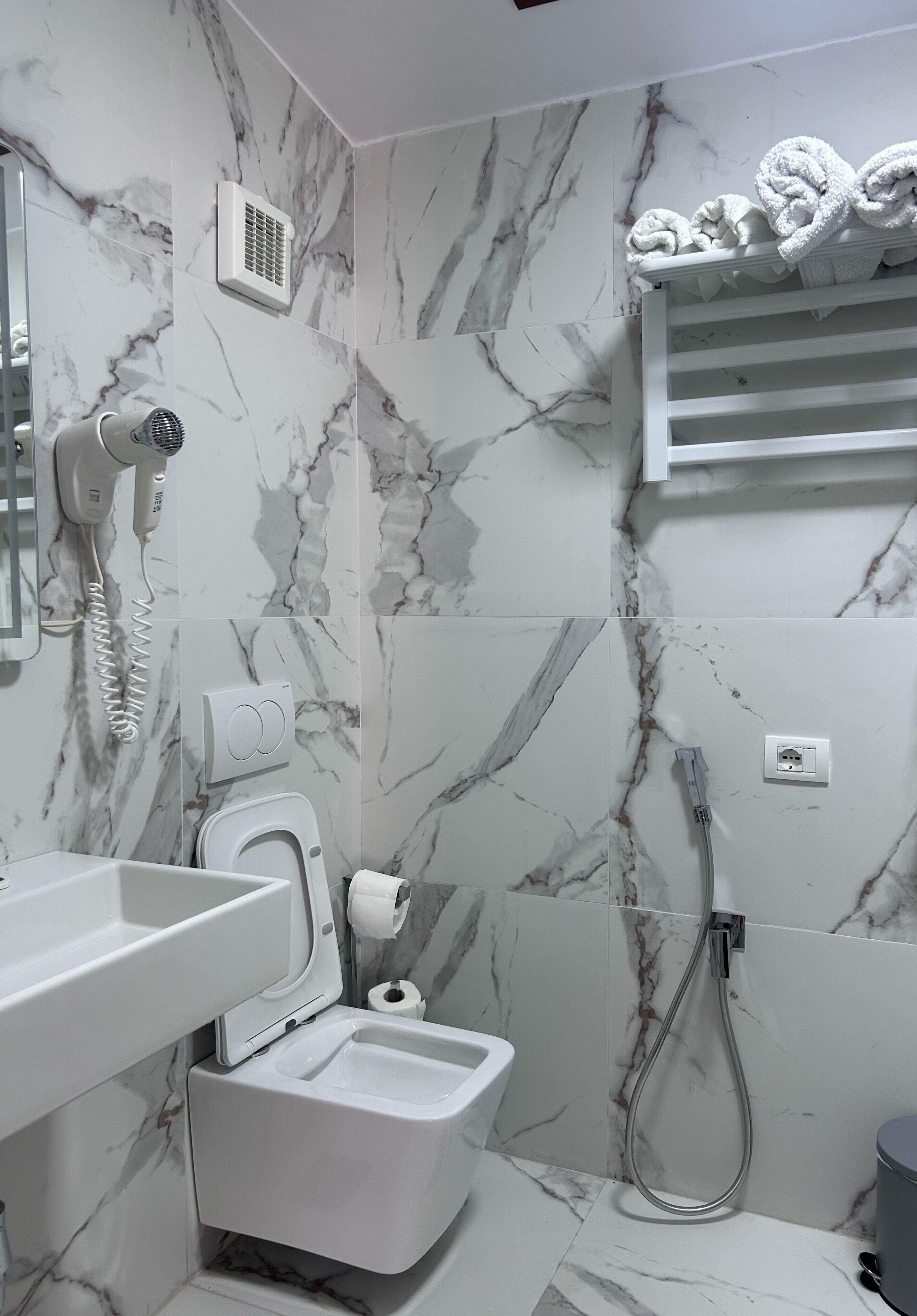 A bathroom with a marble wall, a toilet, a sink, a towel rack, and a shower.