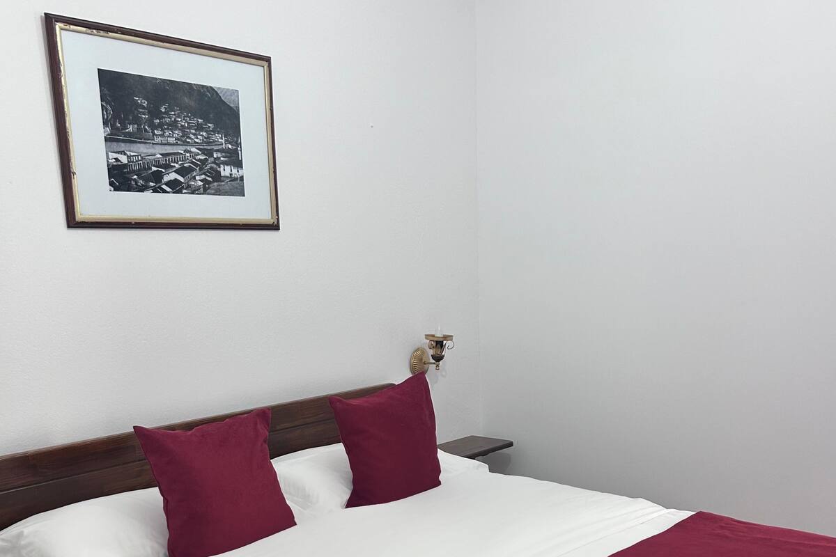 A neatly made bed with white and maroon bedding, a framed picture on the wall, and a patterned rug on the floor.