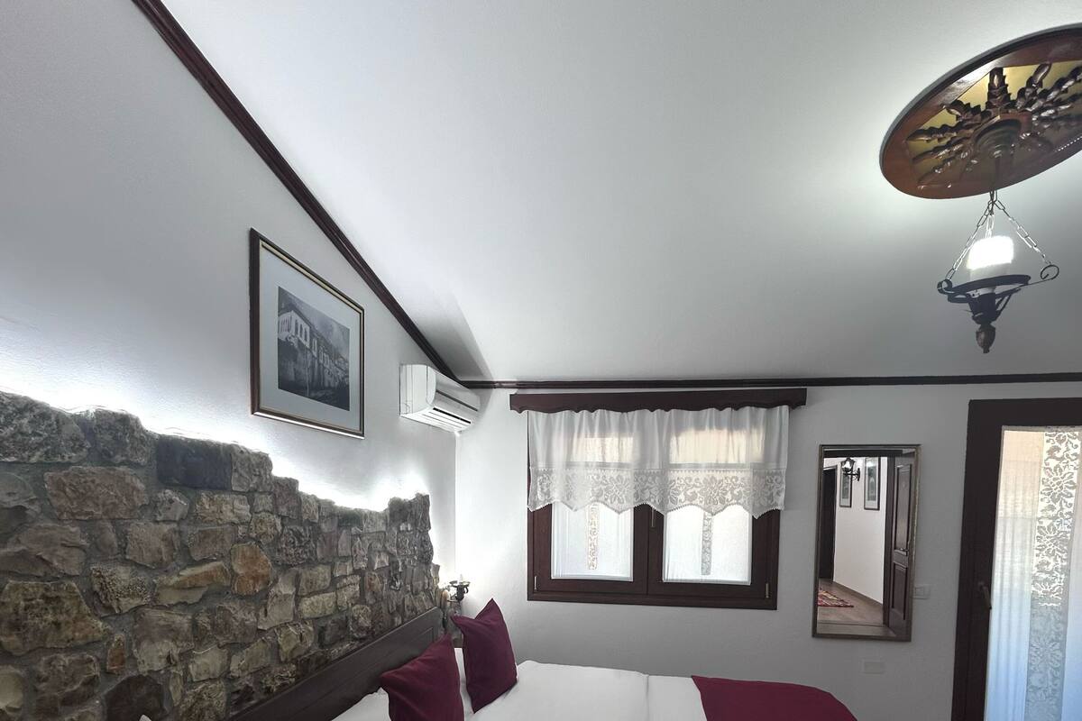 A bedroom with a stone wall, a bed with white and maroon bedding, a wooden headboard, a nightstand, a wall-mounted air conditioner, a window with lace curtains, a mirror, and a ceiling light fixture.