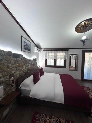 A bedroom with a stone wall, a bed with white and maroon bedding, a wooden headboard, a nightstand, a wall-mounted air conditioner, a window with lace curtains, a mirror, and a ceiling light fixture. - Hotel Berati (Berat)