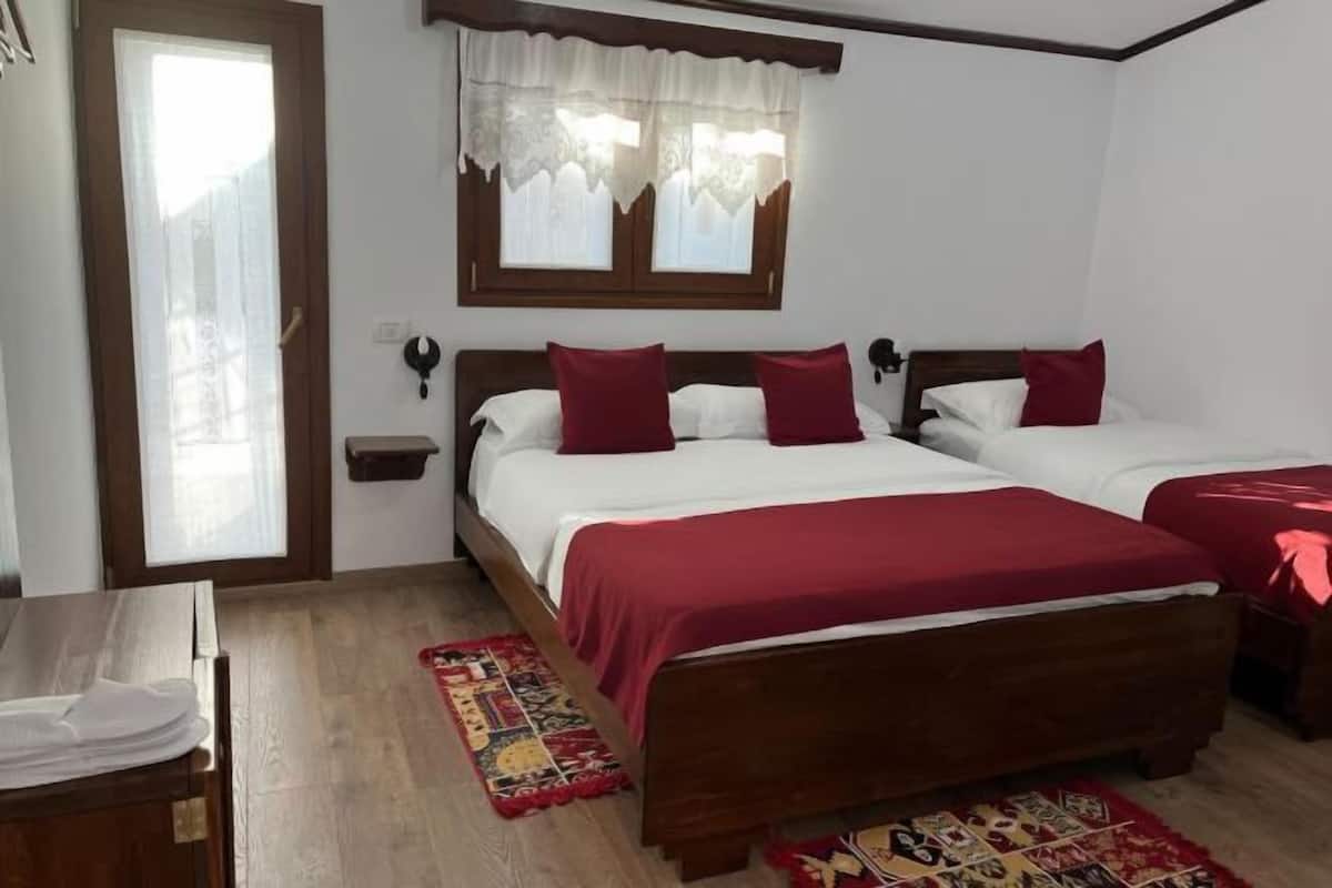 A bedroom with two beds, a wooden headboard, a window with lace curtains, a nightstand, and a red rug.