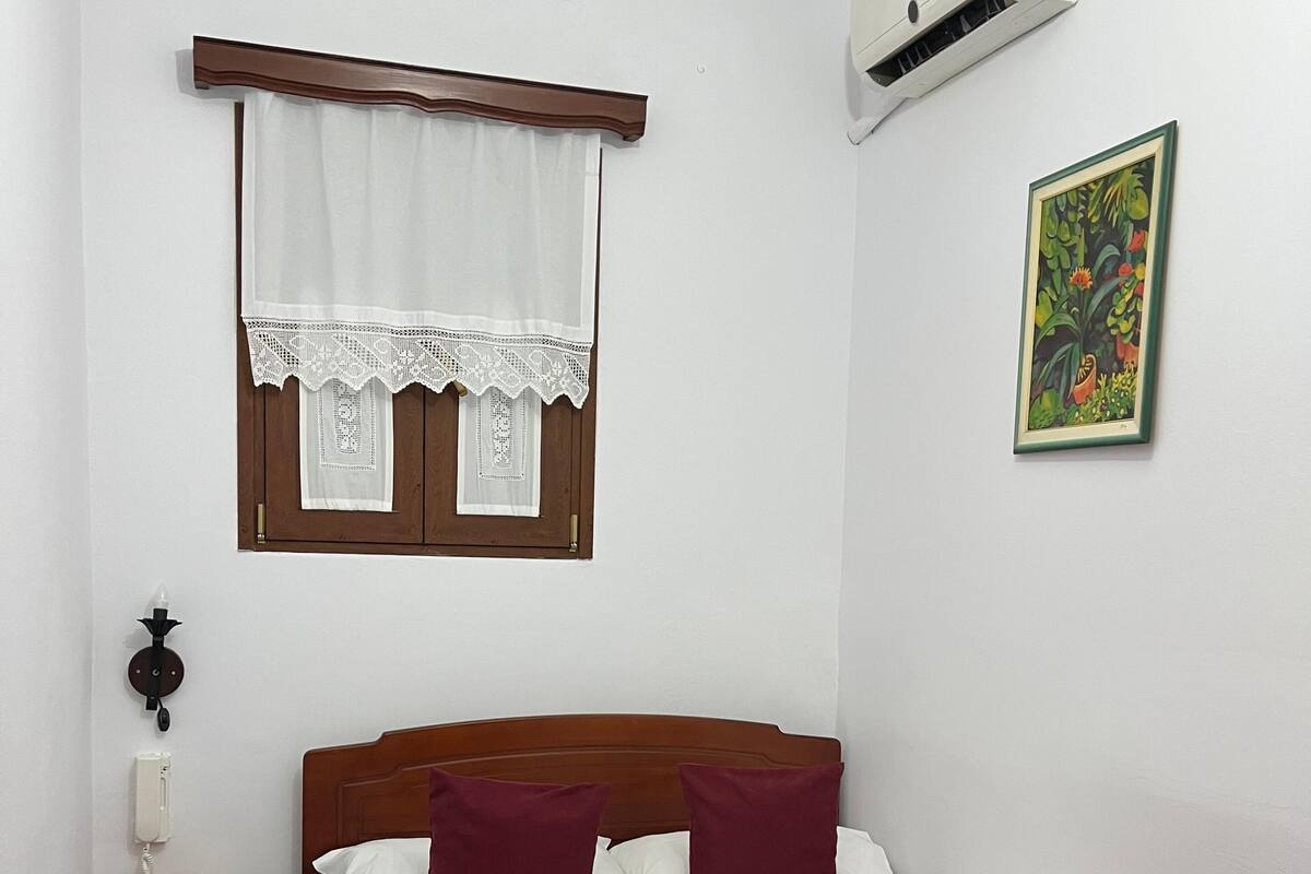 A bedroom with a wooden bed, a window with lace curtains, a wall-mounted air conditioner, a bedside table with a lamp, and a rug on the floor.