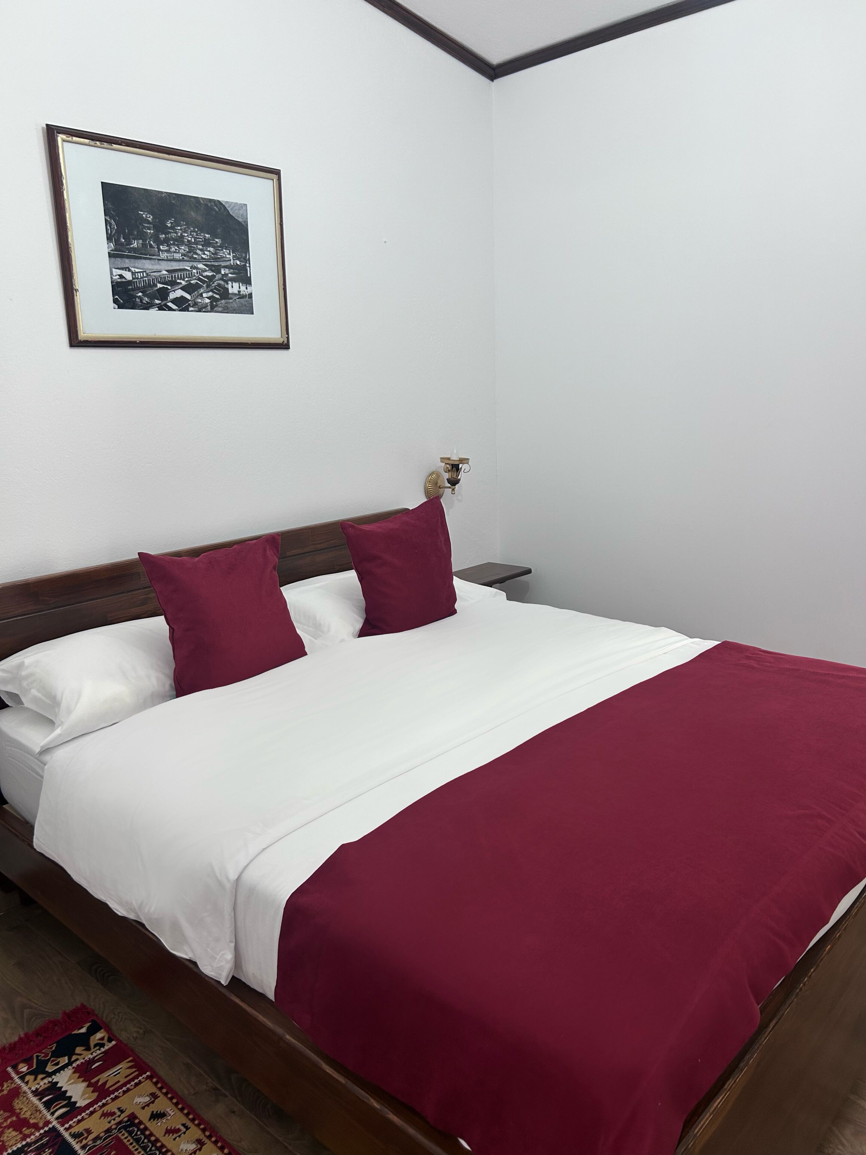 A neatly made bed with white and maroon bedding, a framed picture on the wall, and a patterned rug on the floor.