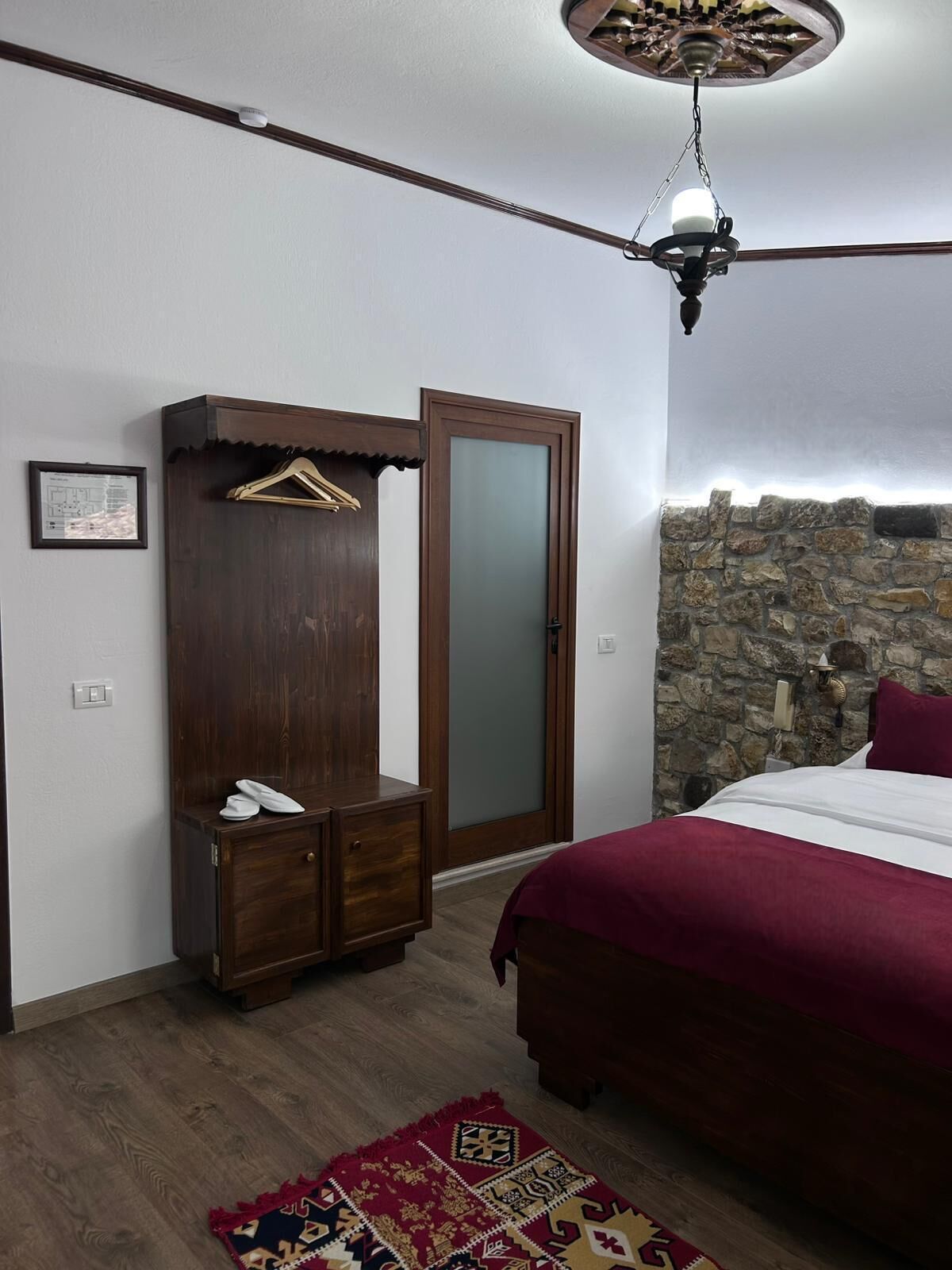 A bedroom with a bed, a wardrobe, a hanging rack, a ceiling fan, a stone wall, and a patterned rug.