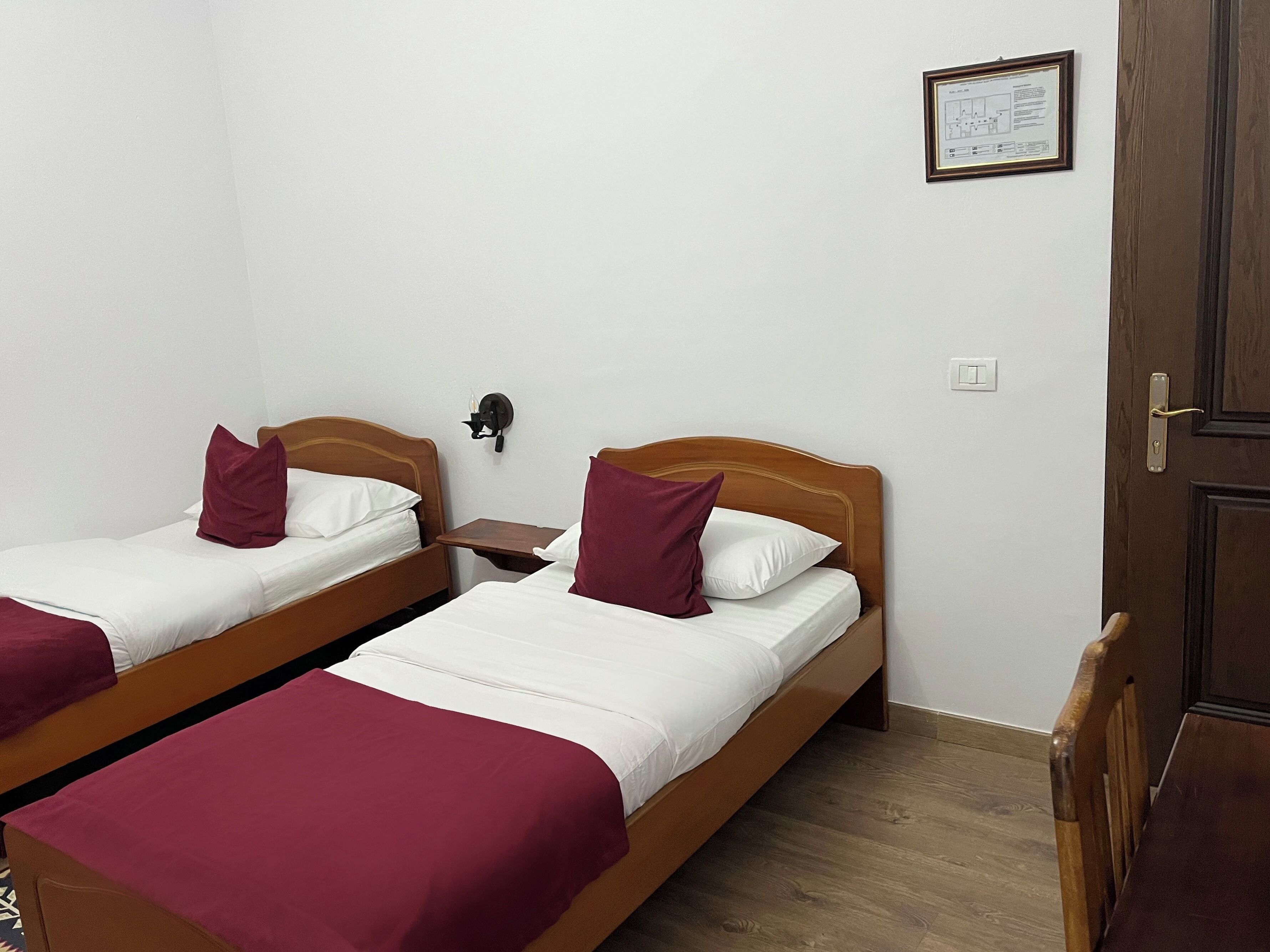 A room with two wooden beds, each with a white mattress and a red blanket. There is a small bedside table with a lamp, a framed certificate on the wall, and a wooden door.