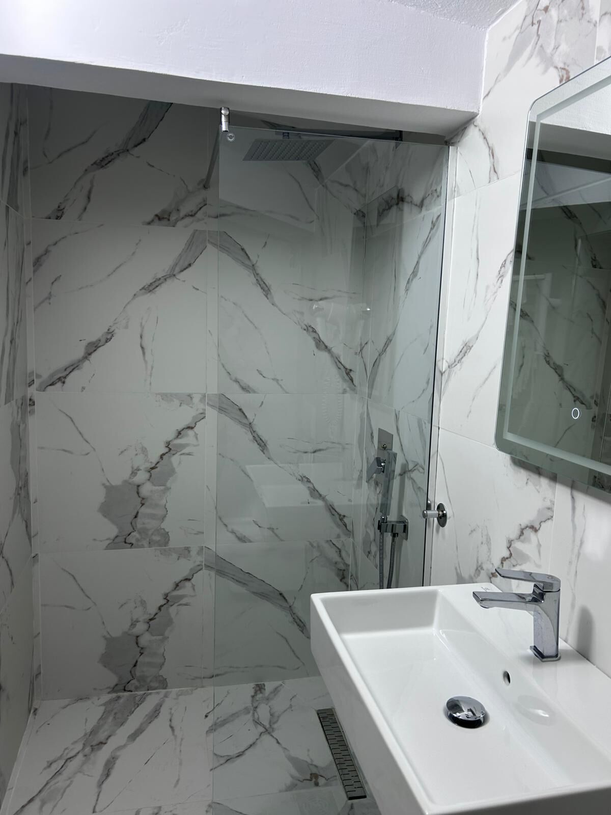 Un bany amb dutxa tancada per vidre, un lavabo blanc i una paret amb un disseny de marbre.
