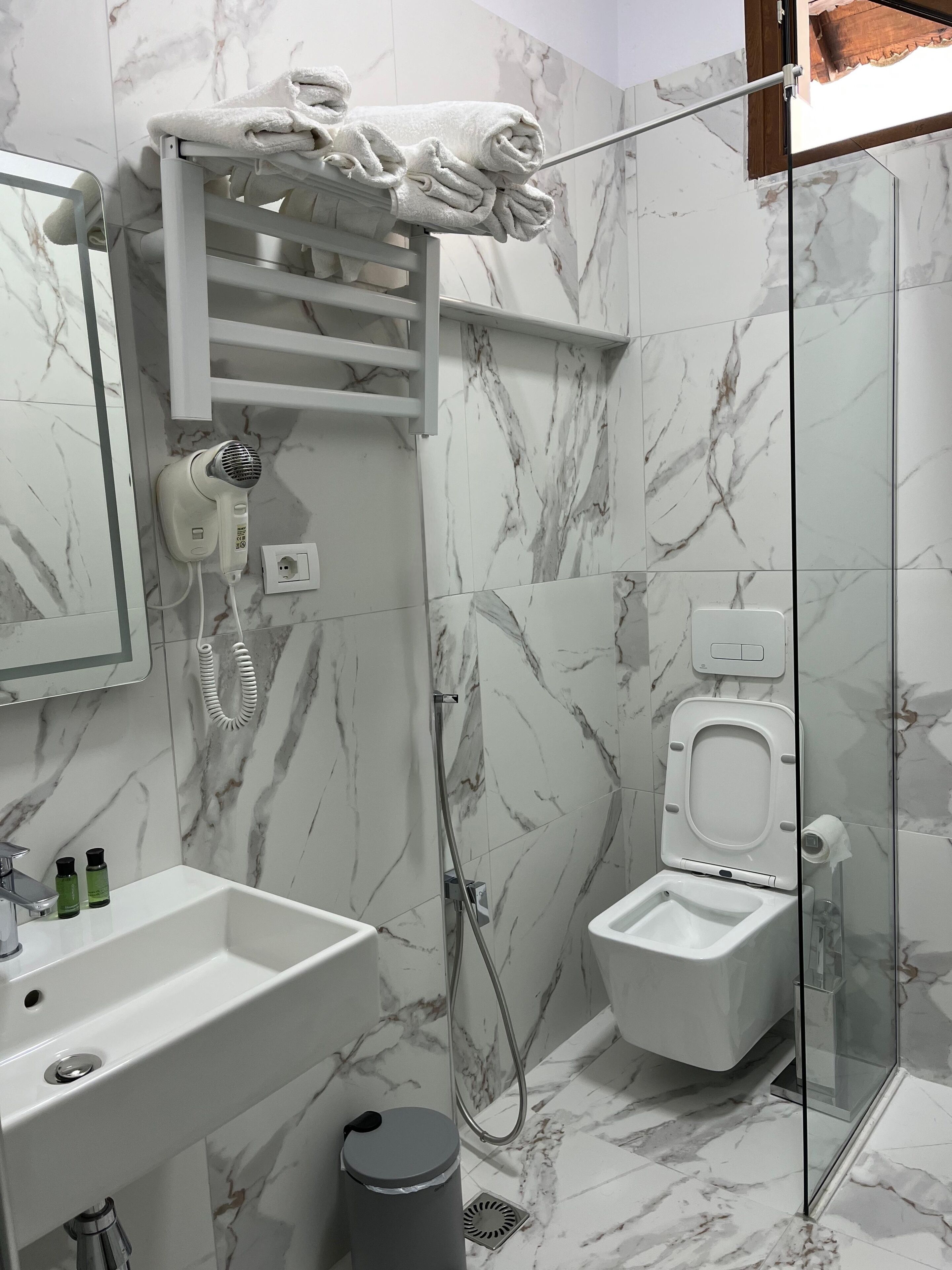 Un baño con pared de mármol, un lavabo blanco, un secador de pelo de mano, cabina de ducha de cristal y un inodoro.