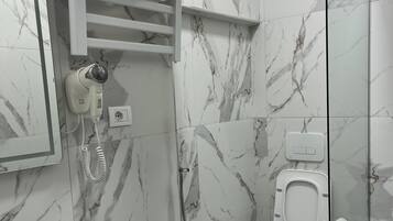 Un baño con pared de mármol, un lavabo blanco, un secador de pelo de mano, cabina de ducha de cristal y un inodoro.