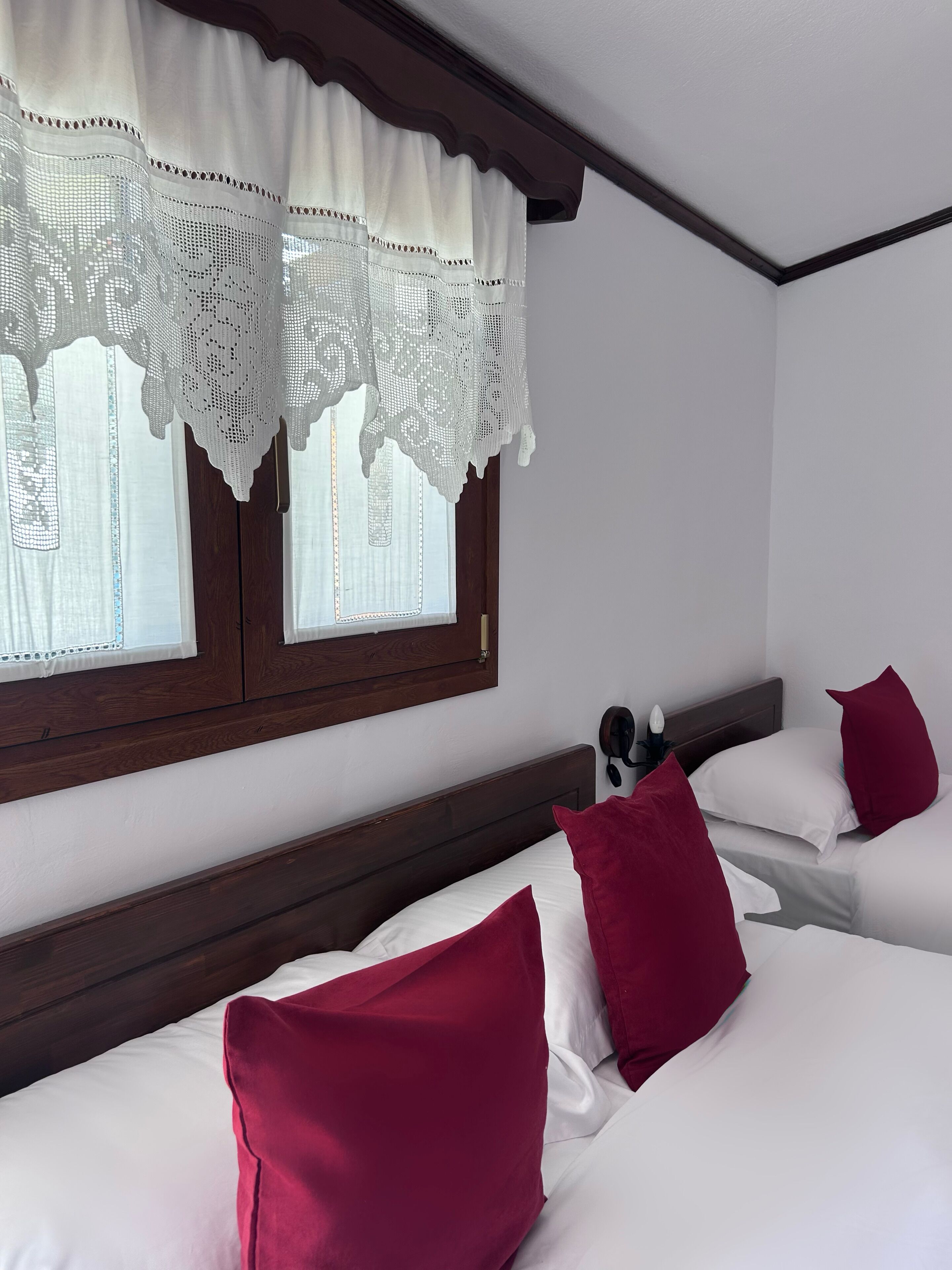 A hotel room with two beds, white bedding, red pillows, a wooden headboard, and a window with lace curtains.
