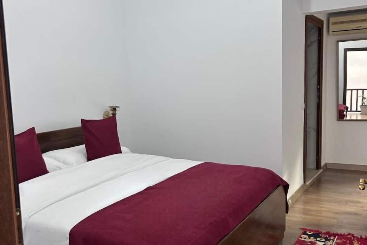 A bedroom with a bed, a wooden headboard, a red throw blanket, and a small bedside table.