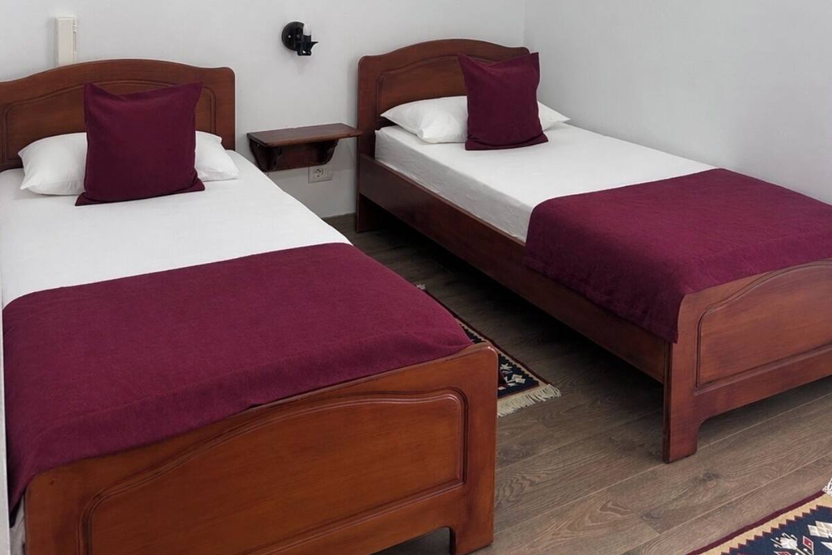 A room with two wooden beds, each with a white mattress and a purple blanket. There is a framed picture on the wall above the beds.