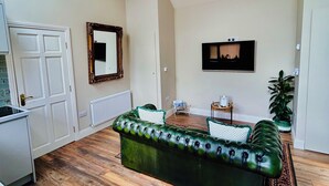Un salon spacieux avec un canapé en cuir vert, un téléviseur fixé au mur et une plante en pot.
