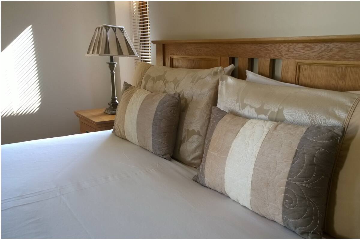 A neatly made bed with a wooden headboard, a bedside table with a lamp, and a window with blinds.