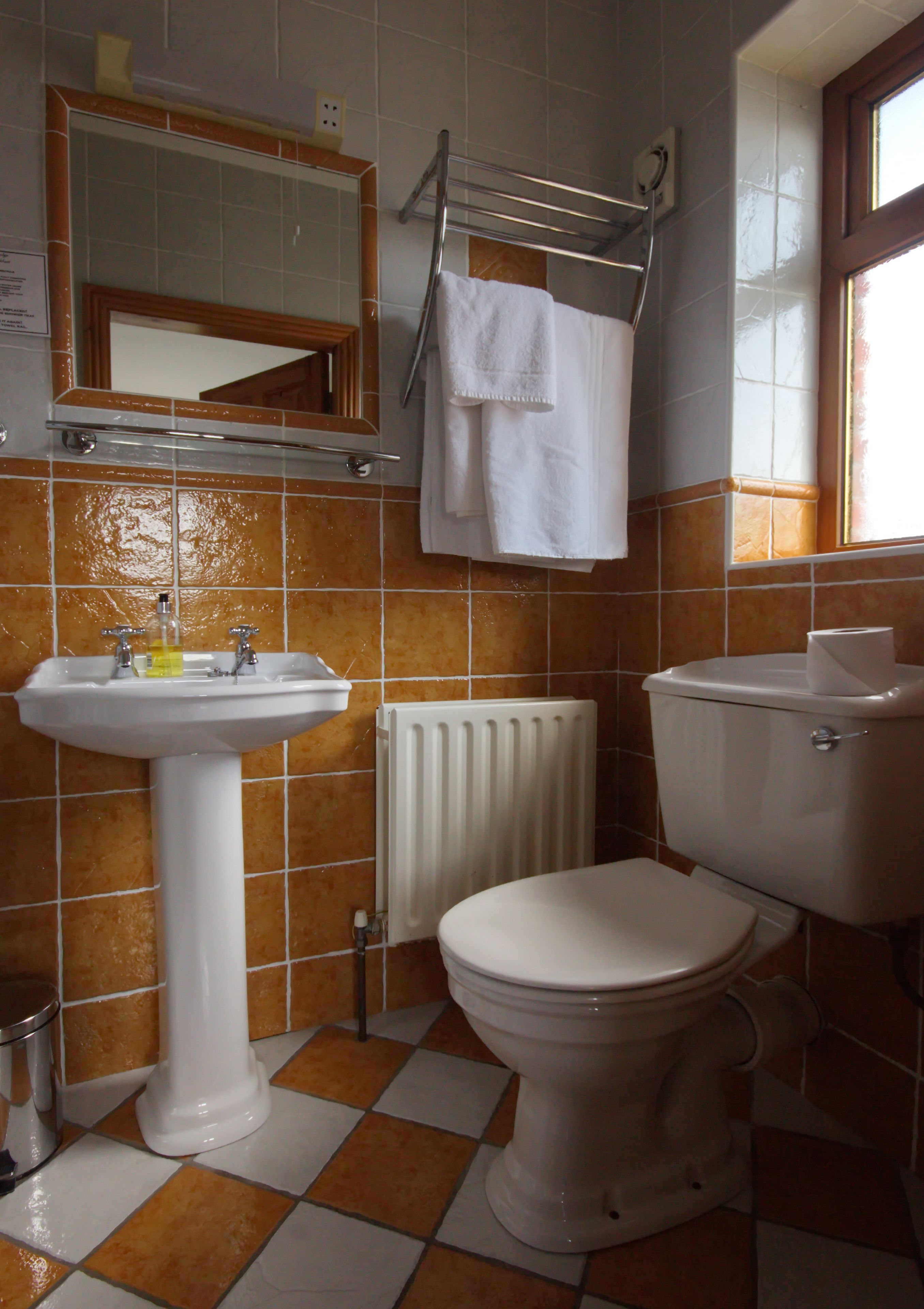 A bathroom with a white sink, toilet, and mirror. There is a towel rack with a white towel hanging on it. The floor has a checkered pattern of orange and white tiles.