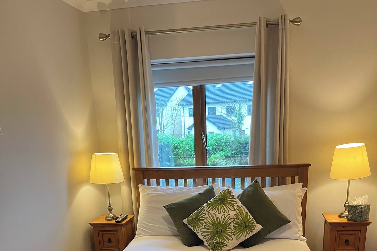 A neatly made bed with a green patterned blanket, two bedside lamps, and a window with curtains.