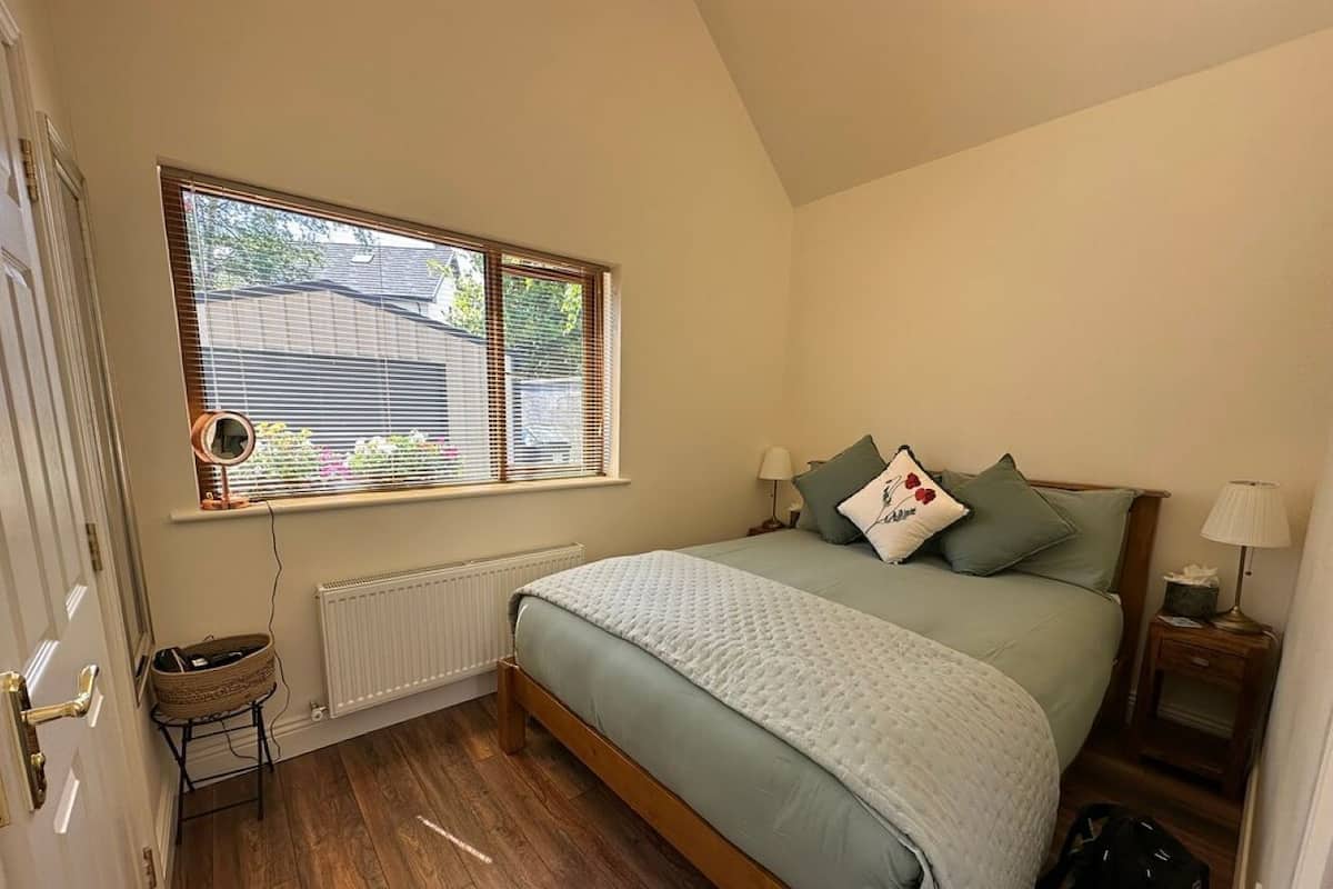 A bedroom with a bed, a window with blinds, a radiator, a bedside table with a lamp, and a small stool.