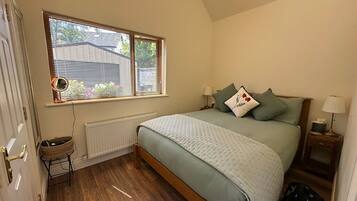 Een slaapkamer met een bed, een raam met zonwering, een radiator, een nachtkastje met een lamp en een klein krukje.