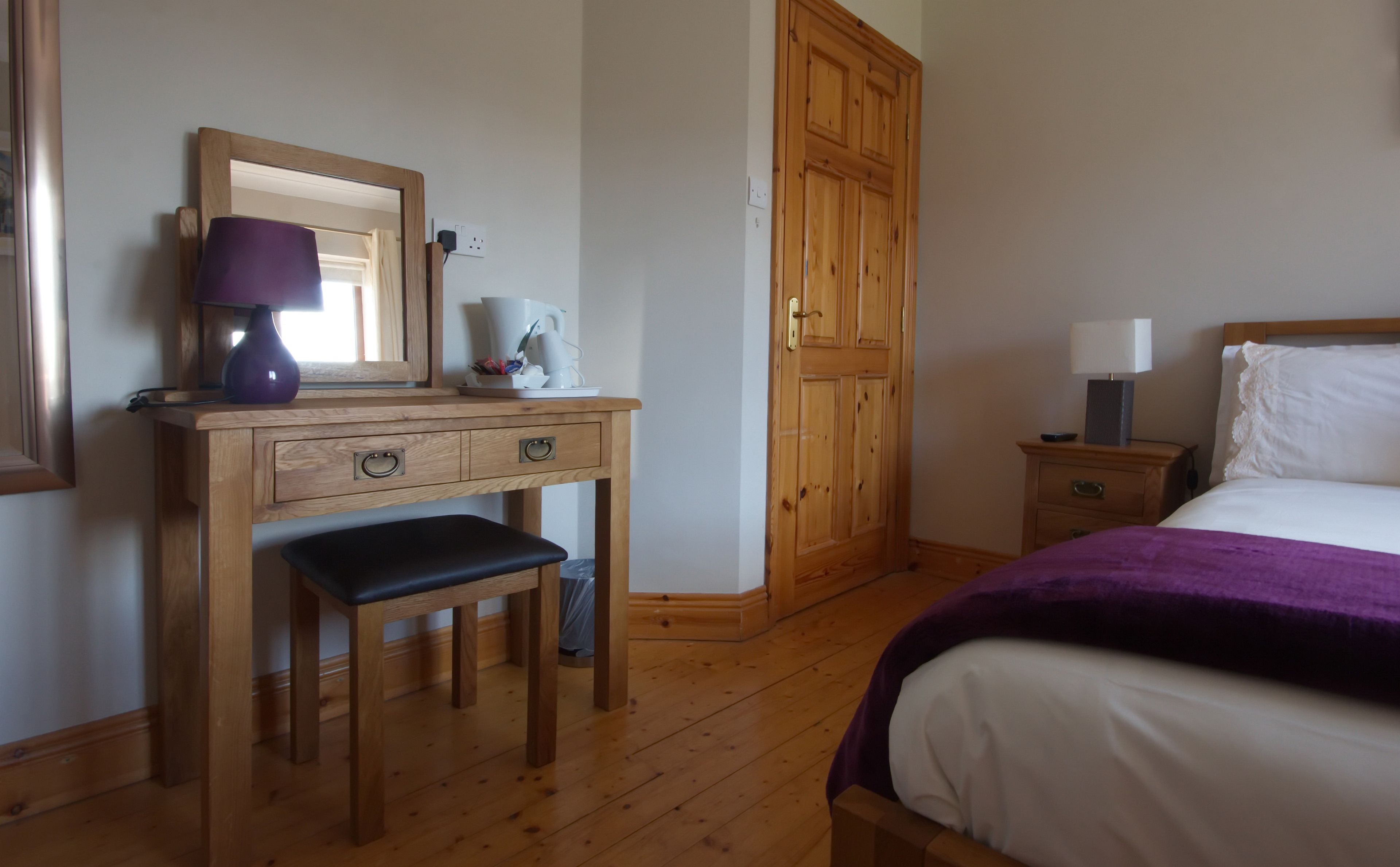 Ein Schlafzimmer mit einem hölzernen Schminktisch, einer Bank, einem Bett mit violetter Decke, einem Nachttisch, einer Lampe und einem Kleiderschrank.
