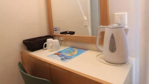 Habitación de hotel con un escritorio de madera, una silla verde, una tetera blanca, una taza y una revista.