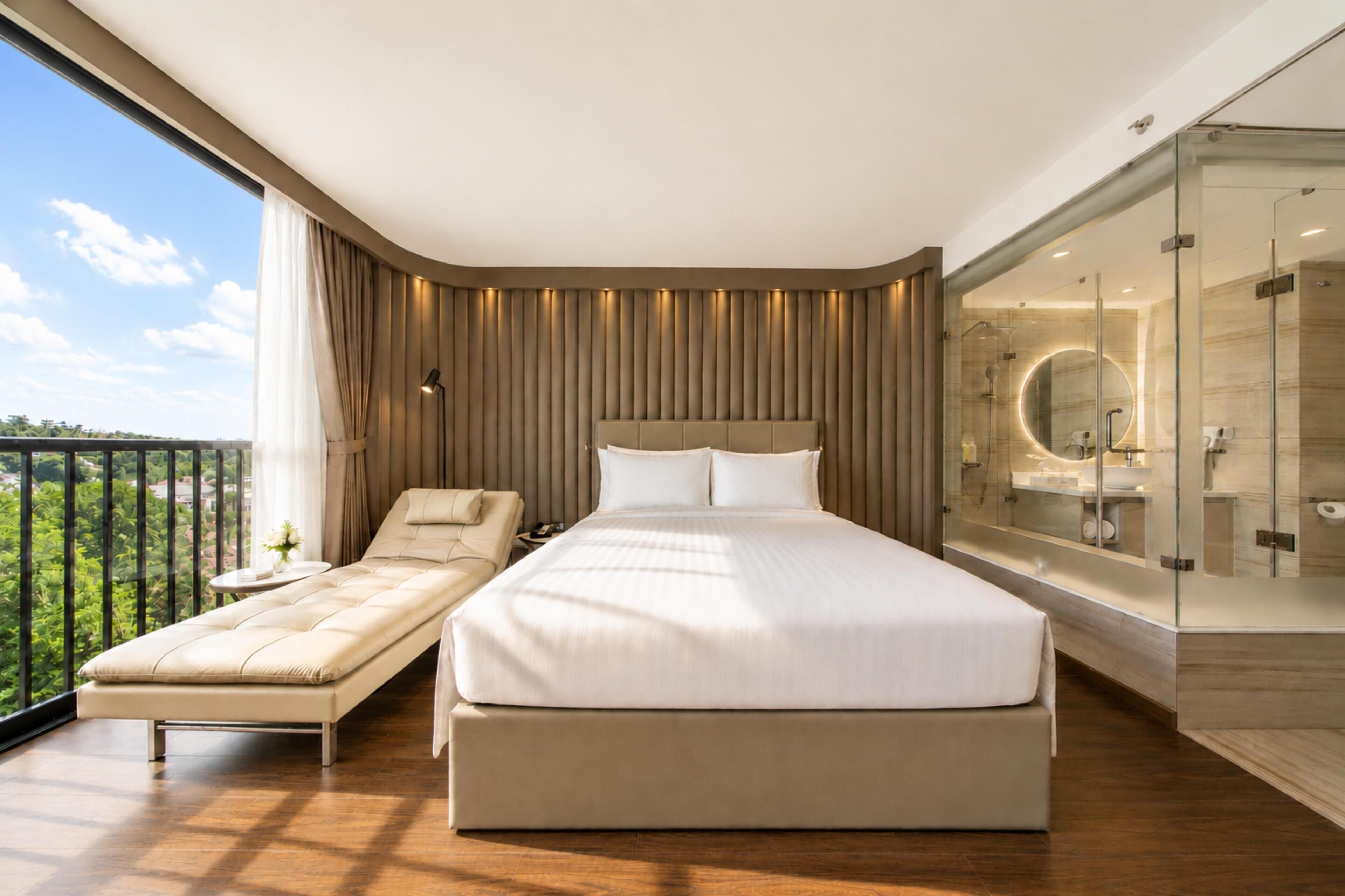 A modern bedroom with a large bed, a chaise lounge, a glass-enclosed bathroom, and a balcony with a view.