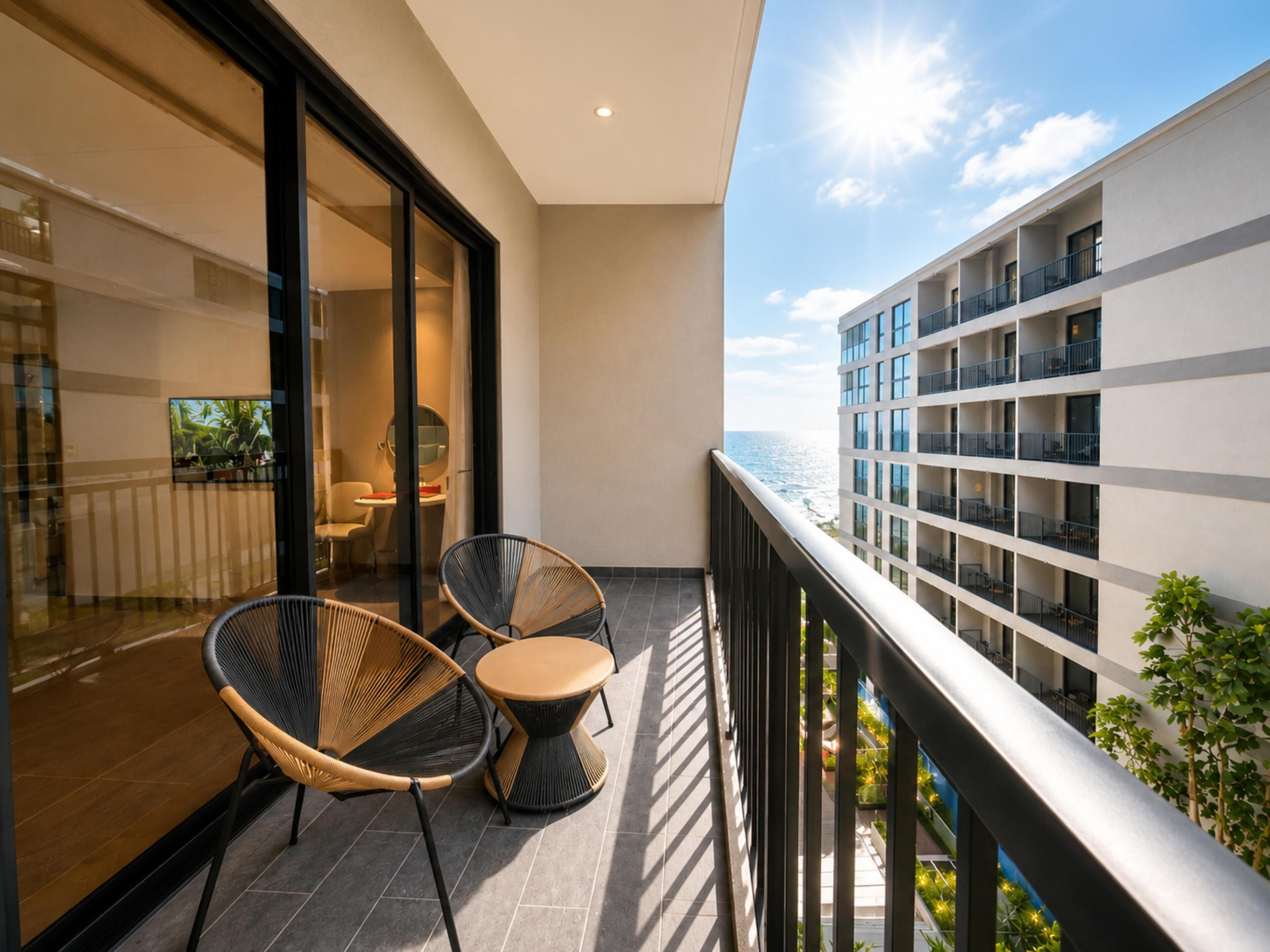 A balcony with two wicker chairs and a small table, overlooking a building with multiple balconies and a view of the sea.