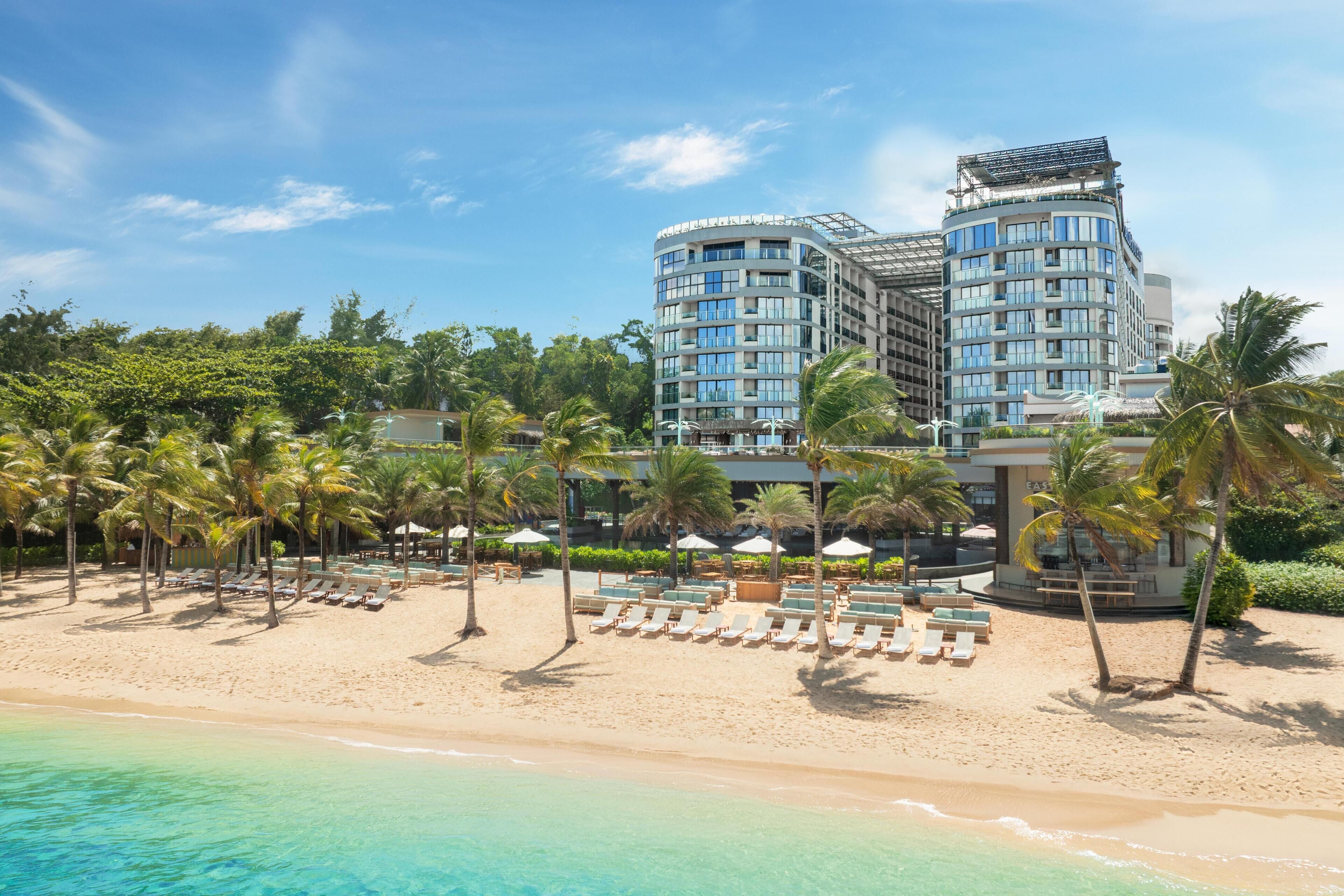 A beachfront hotel with multiple floors, a sandy beach, and palm trees.