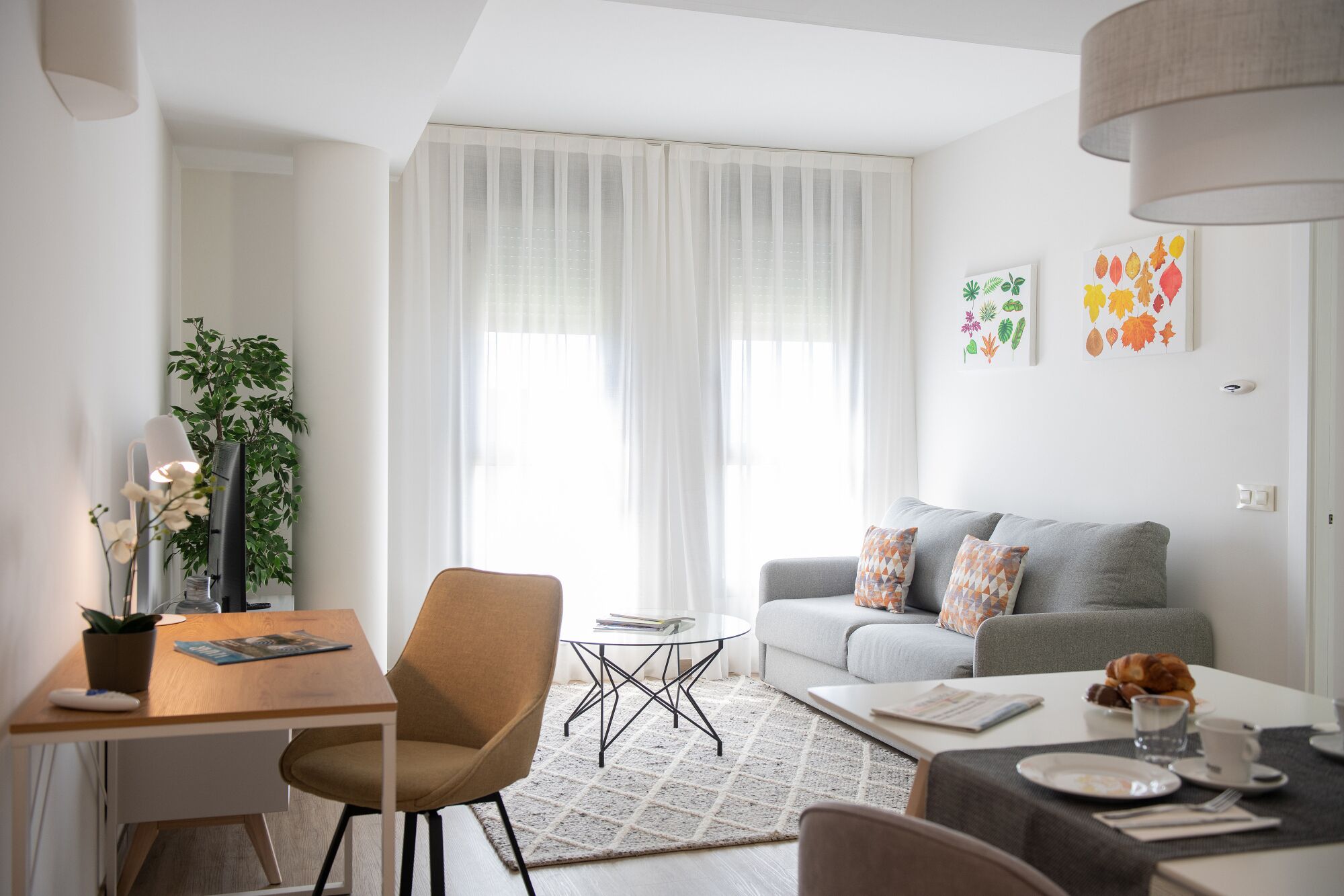 Ein modernes Wohnzimmer mit grauem Sofa, einem Holzesstisch und einem Couchtisch. Links steht eine Pflanze und rechts eine Lampe.