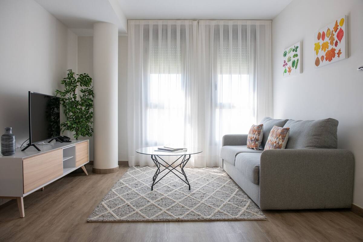 Een moderne woonkamer met een grijze bank, een glazen salontafel, een televisie op een houten dressoir en een groot raam met dunne gordijnen.