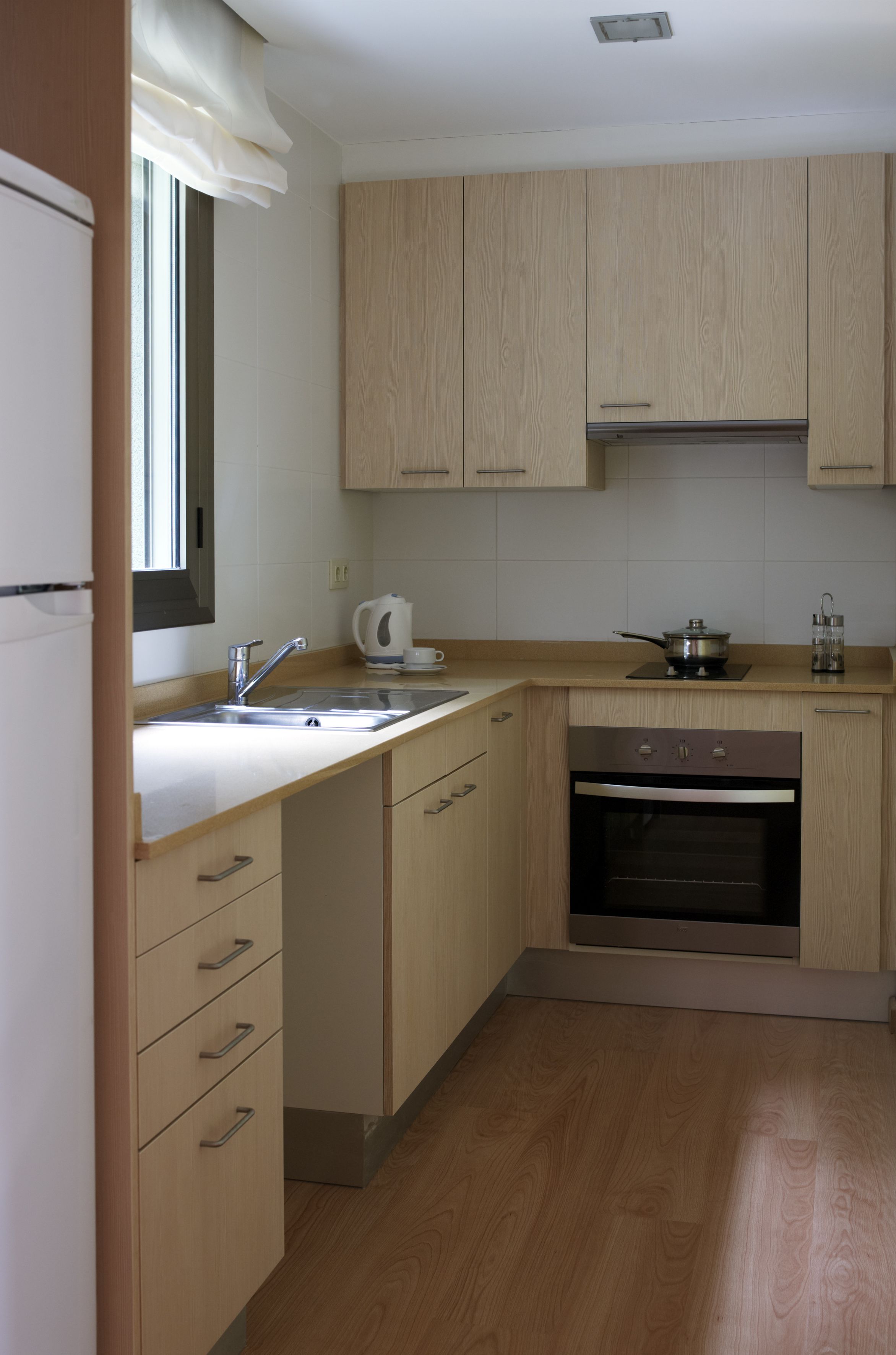 A modern kitchen with wooden cabinets, a stainless steel sink, a built-in oven, and a window with a white valance.
