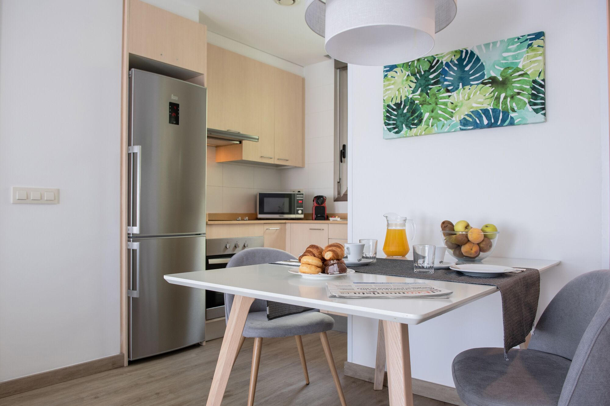 Une cuisine moderne équipée d’un réfrigérateur en acier inoxydable, d’un four à micro-ondes et d’une machine à café. On y trouve une table à manger avec deux chaises, un saladier rempli de fruits et une carafe de jus d’orange.