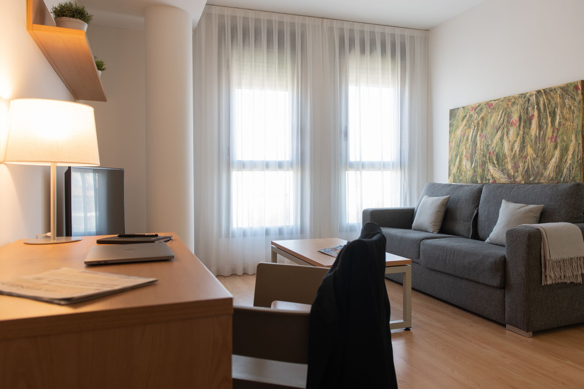 Ein modernes Hotelzimmer mit Schreibtisch, Stuhl, Sofa und großen Fenstern mit transparenten Vorhängen.
