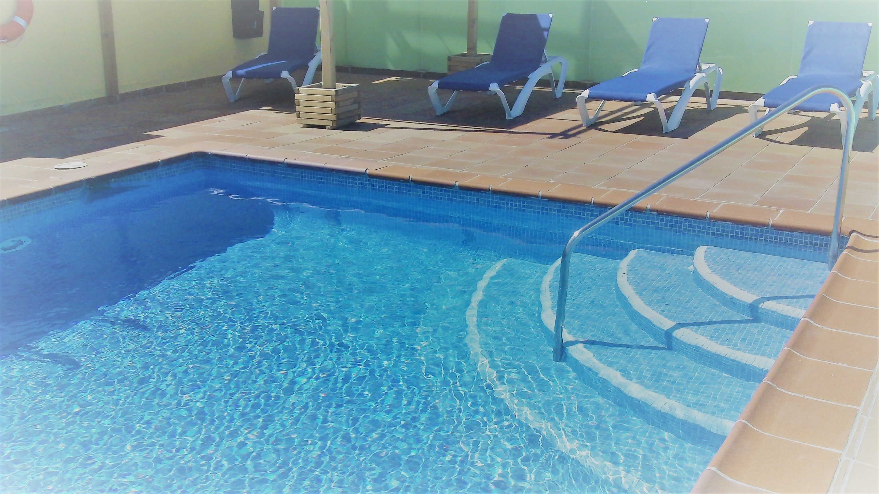 Une piscine rectangulaire à l’eau d’un bleu limpide, entourée d’une terrasse carrelée. On y trouve des chaises longues et un tabouret en bois près du bord de la piscine.