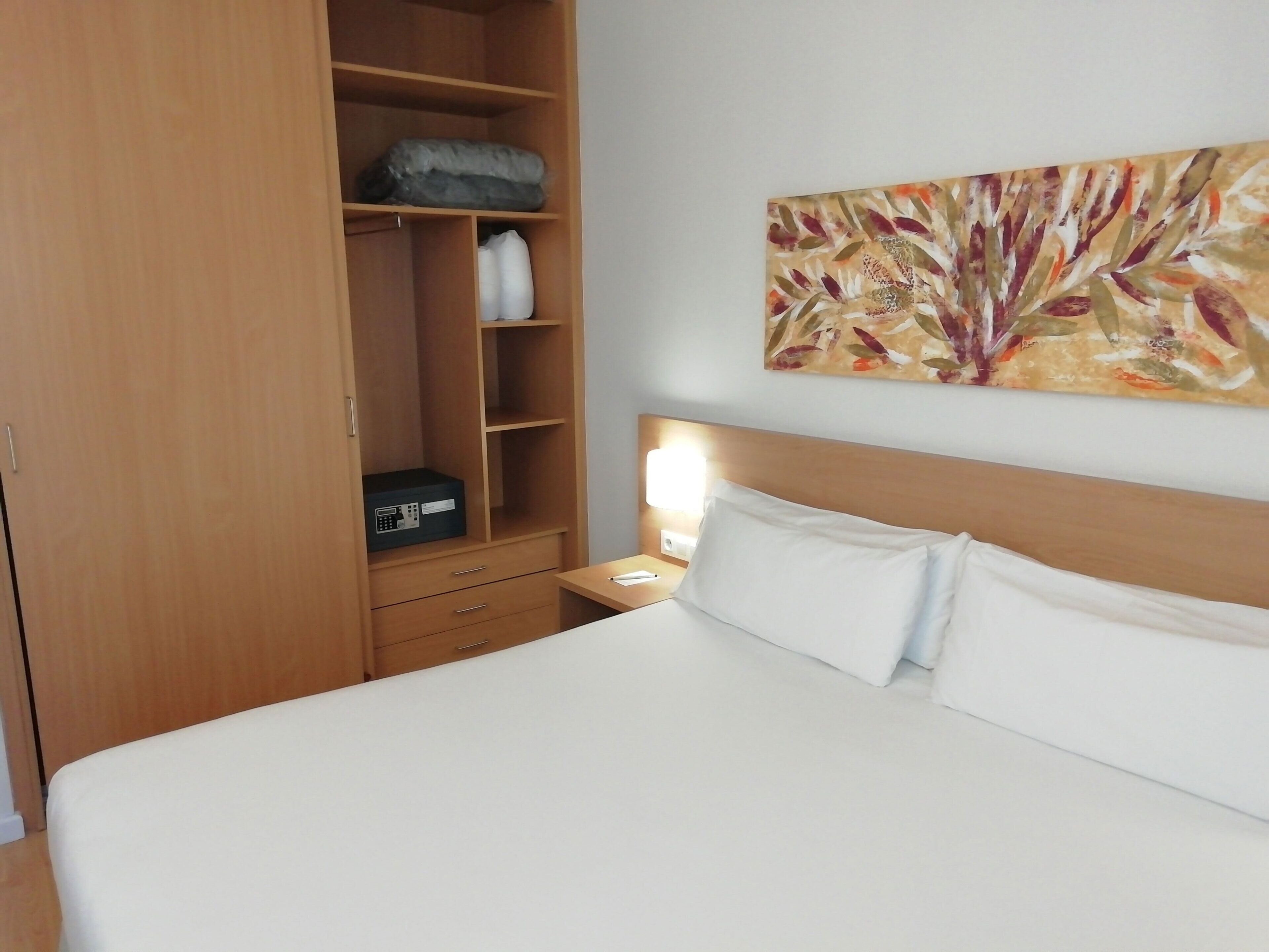 A hotel room with a bed, a wooden wardrobe, a nightstand, and a wall-mounted painting.