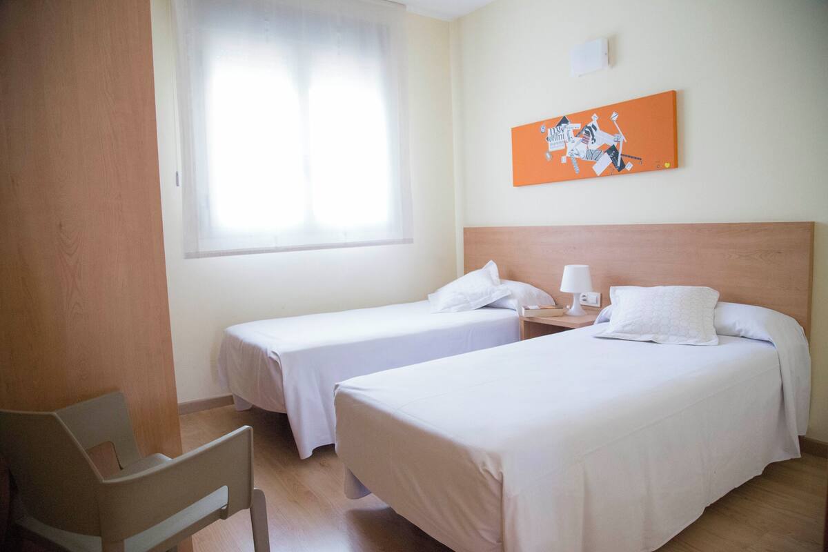 Een hotelkamer met twee enkele bedden, een houten hoofdbord, een nachtkastje met een lamp en een raam met dunne gordijnen.
