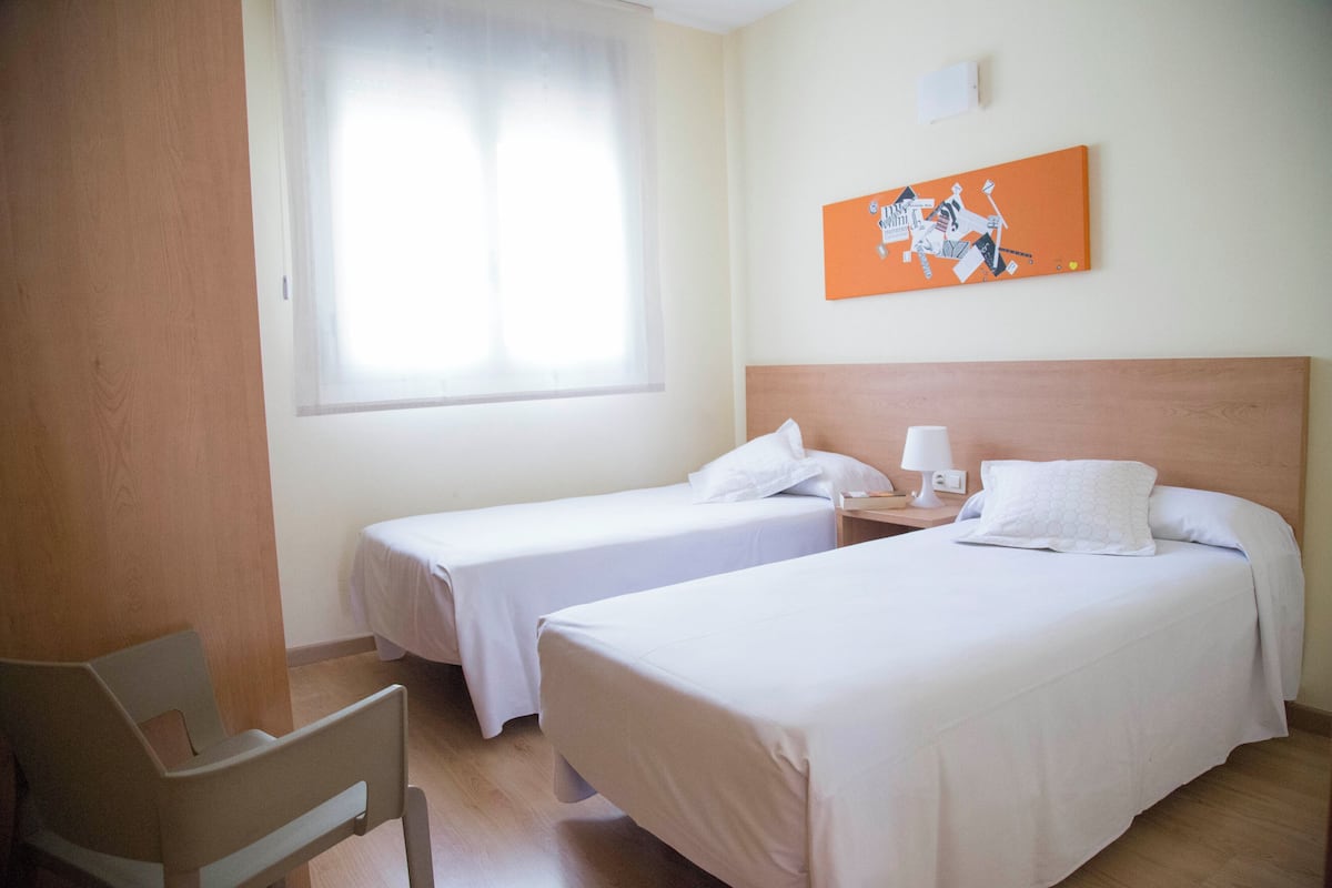 A hotel room with two single beds, a wooden headboard, a nightstand with a lamp, and a window with sheer curtains.