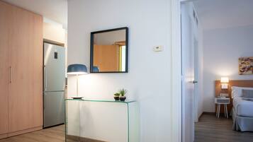 A modern kitchen with a built-in refrigerator, a glass console table with plants, a wall-mounted mirror, and a door leading to a bedroom with a bed and bedside table.