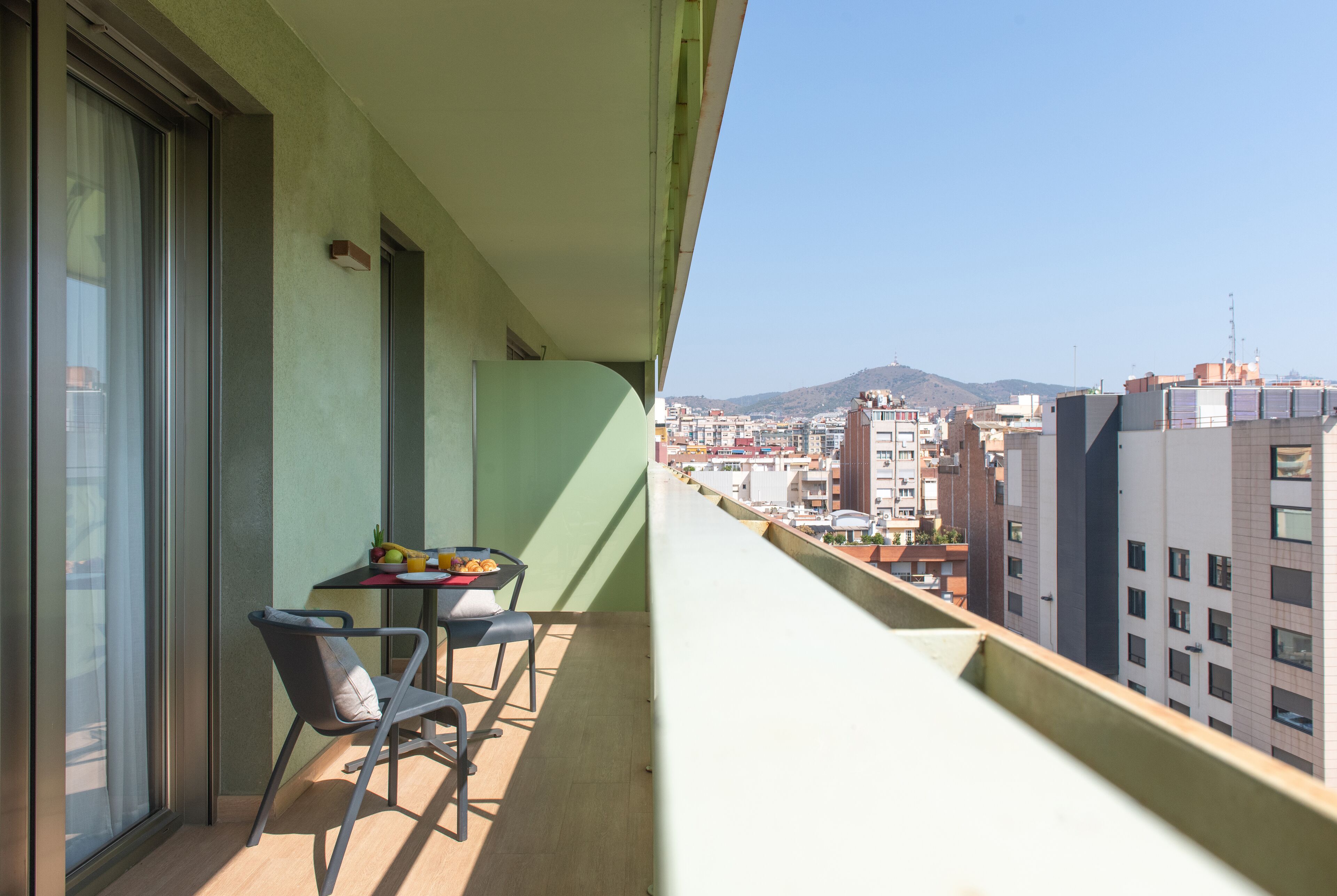 A balcony with a table and chairs, overlooking a cityscape with buildings and a mountain in the distance.