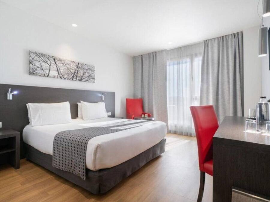 A hotel room with a bed, a desk, a chair, a TV, and a window with curtains.