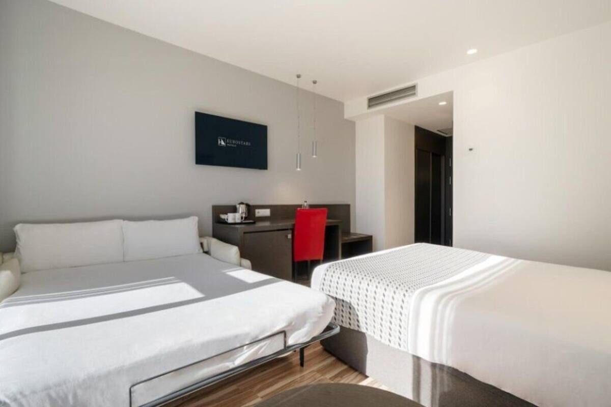 Een hotelkamer met twee bedden, een bureau, een stoel en een televisie aan de muur.