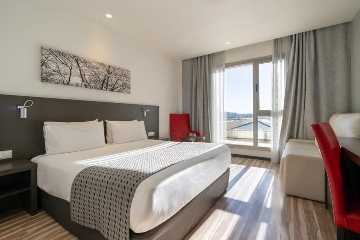 Een moderne hotelkamer met een groot bed, een rode fauteuil, een bureau en uitzicht op de stad door grote ramen.