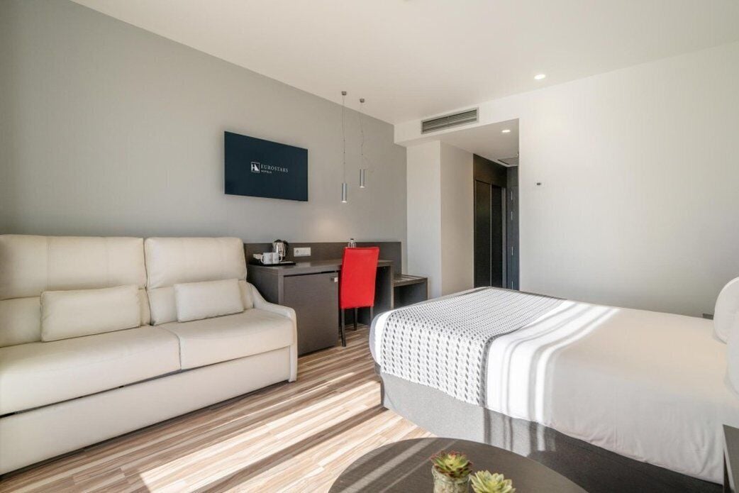 Una habitación de hotel moderna con sofá, cama, escritorio y una silla roja.