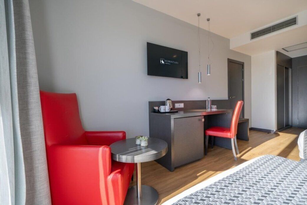 A modern hotel room with a bed, a red sofa, a small round table, a kitchenette, and a TV mounted on the wall.