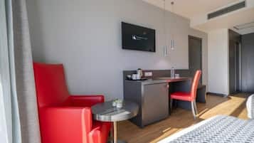 A modern hotel room with a bed, a red sofa, a small round table, a kitchenette, and a TV mounted on the wall.