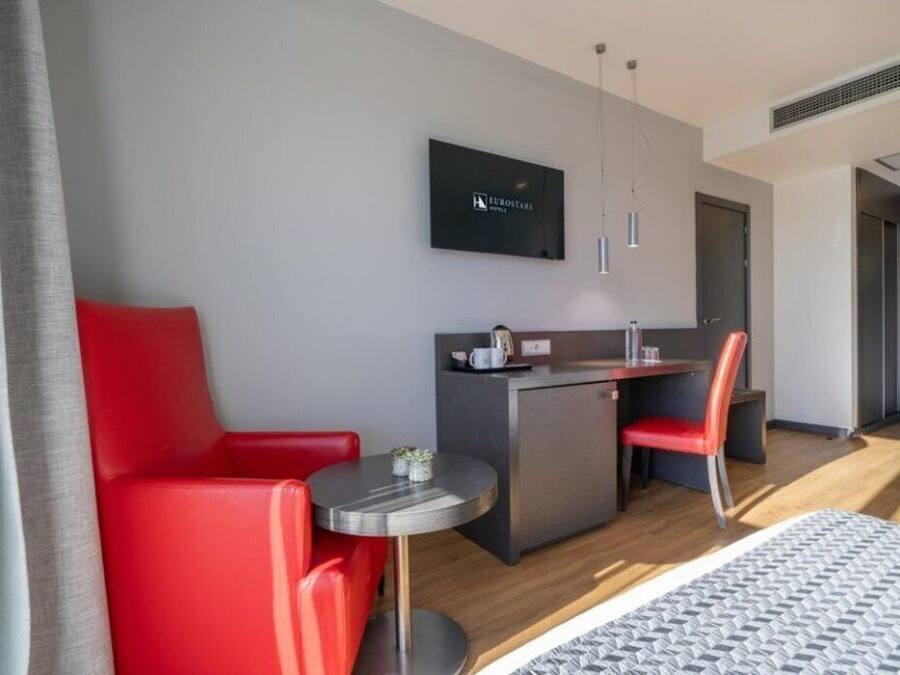 A modern hotel room with a bed, a red sofa, a small round table, a kitchenette, and a TV mounted on the wall.