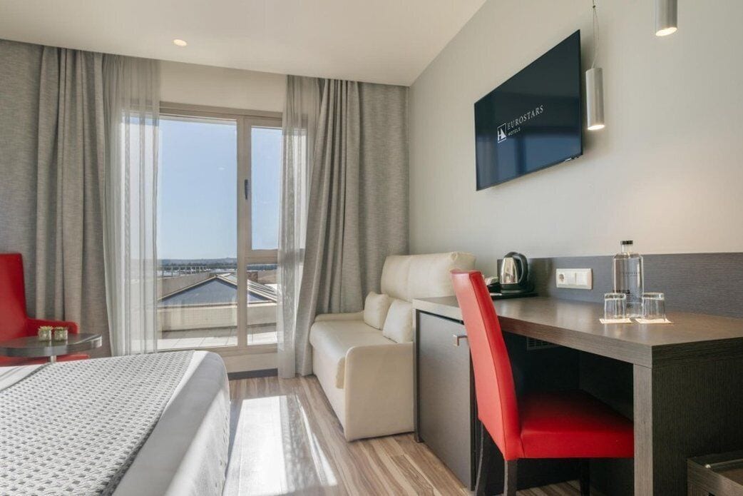 A hotel room with a bed, a desk, a chair, a TV, and a view of the city.