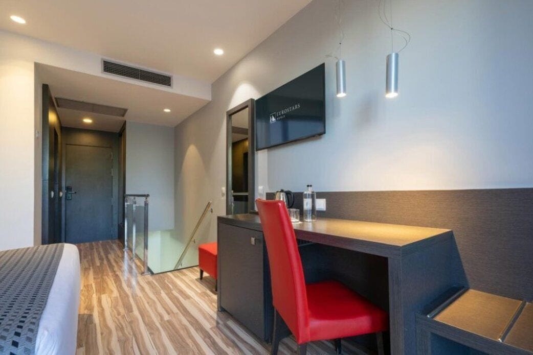 A modern hotel room with a wooden floor, a bed, a desk with a red chair, a small kitchenette, and a TV mounted on the wall.